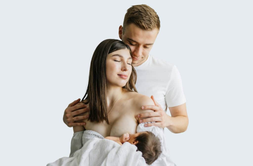 A breastfeeding mom is holding a newborn baby and a man is hugging her.