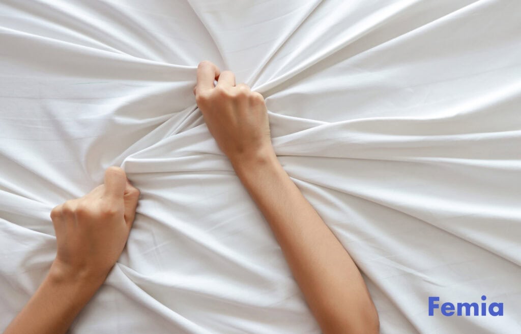 A woman gripping a bedsheet with both hands illustrates a breastfeeding orgasm.