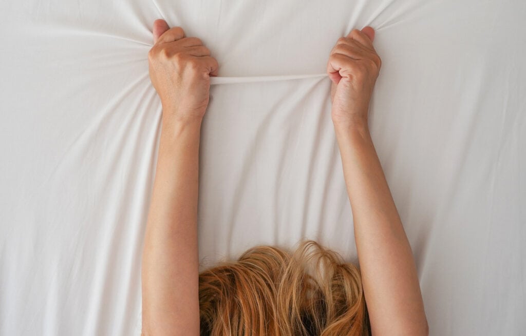 A woman gripping a bedsheet with both hands, illustrating an orgasm during pregnancy.