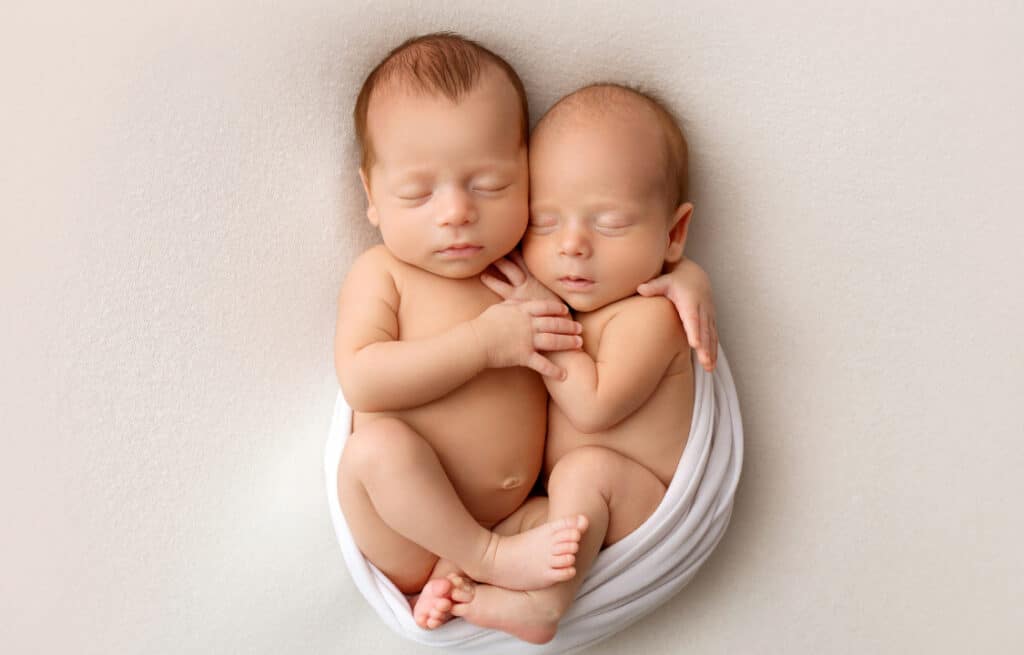 Newborn twins sleeping together wrapped in a white blanket.