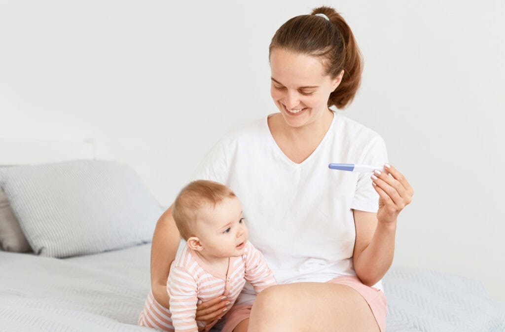 A picture of a woman holding a baby and a positive pregnancy test, representing pregnancy while breastfeeding.