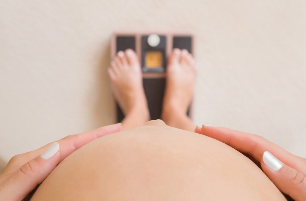 A picture of a pregnant woman on a bathroom scale holding her belly.