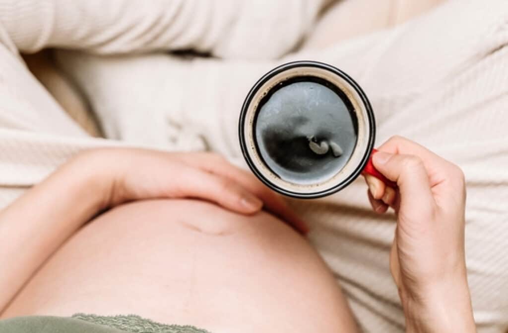 A photo of a baby bump with a cup of coffee representing the question ‘Can I drink coffee while pregnant?’.