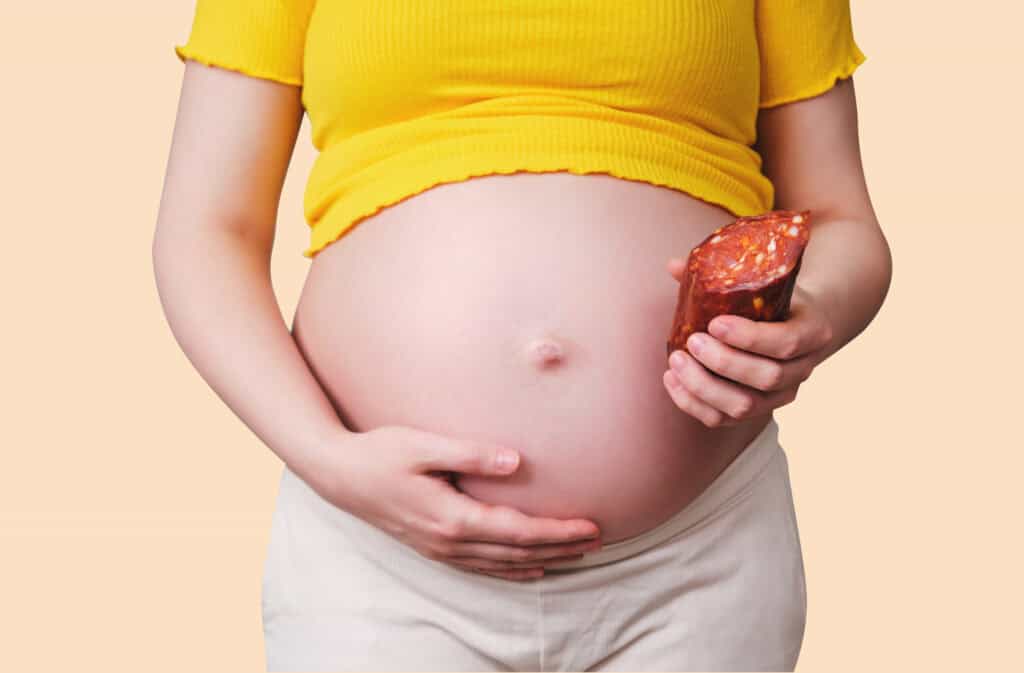 A photo of a pregnant woman, holding a belly in one hand and a deli meat in another.