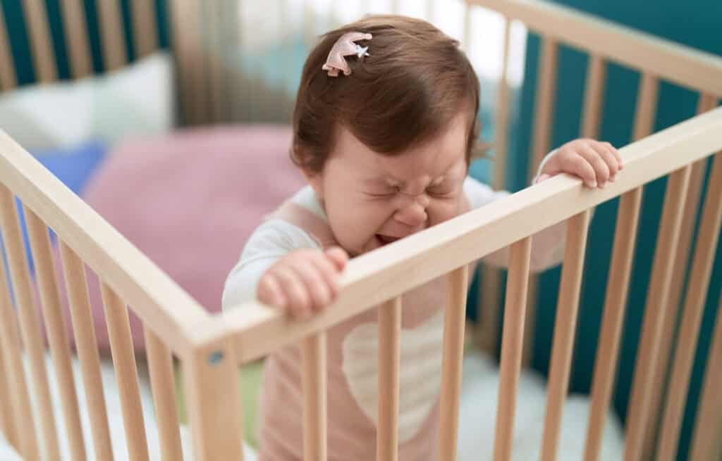 A photo of a crying baby standing in a crib, experiencing 9-month sleep regression.