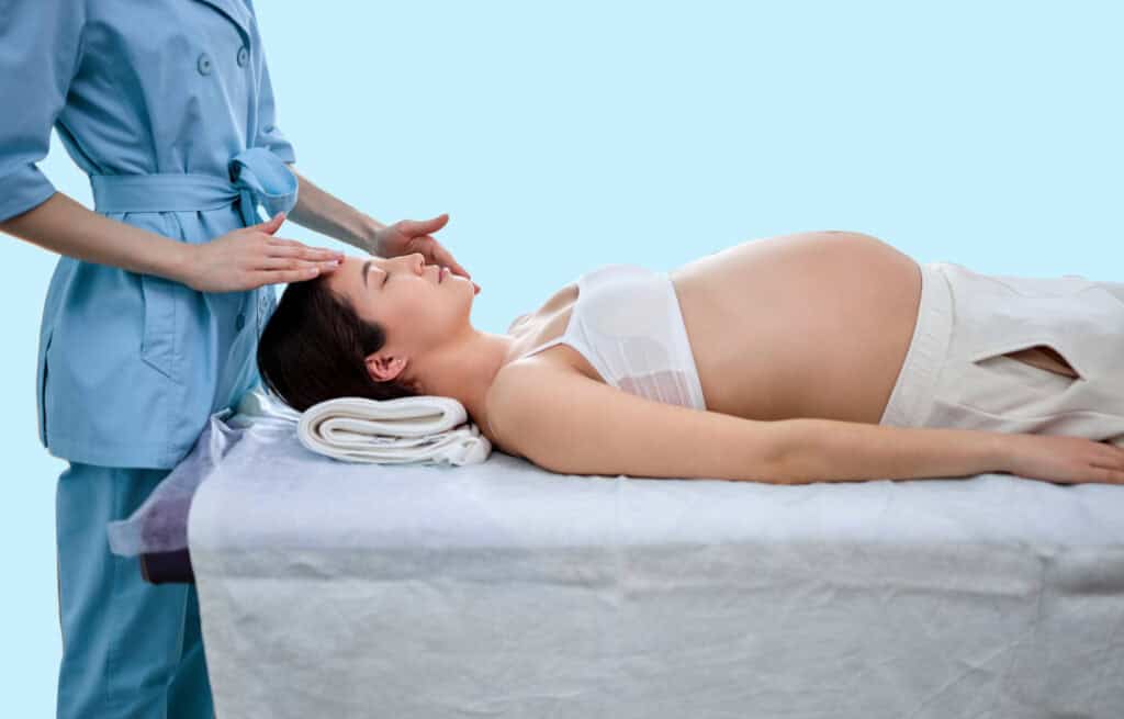 A photo of a pregnant woman laying, and the esthetician over her, wondering 'can you get a facial while pregnant?'