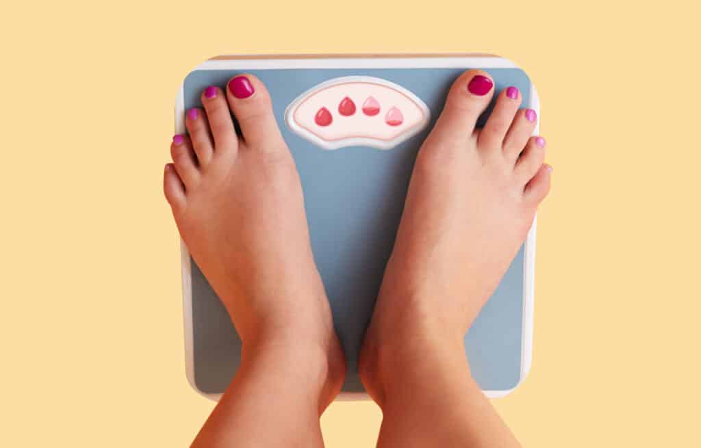 A photo of a woman's feet standing on a scale with red drops instead of numbers, representing the question, 'Do you weigh more on your period?'